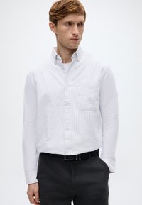 Λευκό πουκάμισο με κουμπιά και διακριτικό ριγέ μοτίβο, μία τσέπη στο στήθος και γιακά. Φορεμένο με σκούρο γκρι παντελόνι και μαύρη ζώνη.