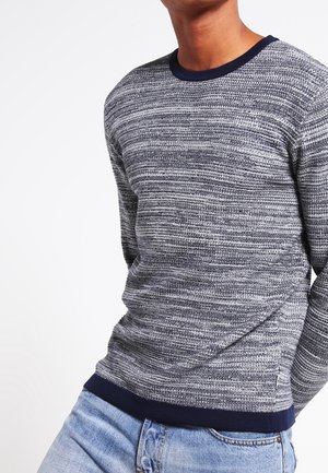 Homme portant un pull en maille rayé gris et bleu marine avec des poignets et un ourlet bleu marine, assorti à un jean bleu clair, sur fond blanc.