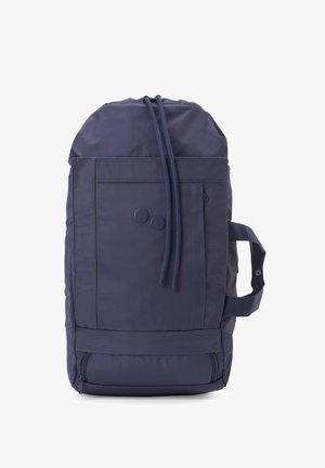 Frau mit einem großen marineblauen Rucksack mit Seitentaschen und einem Reißverschlussfach am Boden, gekleidet in schwarzes Oberteil und graue Hose.