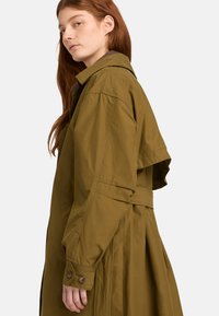 Trench-coat kaki avec une ceinture à la taille, un grand col et un détail à rabat sur les épaules. Comprend des poignets boutonnés et une texture douce.