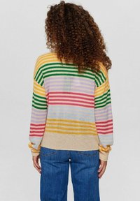 Žena s kudrnatými vlasy je oblečena v pruhovaném svetru v několika barvách a modrých džínách, stojí zády k divákovi na neutrálním pozadí.