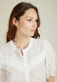 Chemisier blanc avec broderie en œillet, manches courtes à volants, col rond et fermeture à bouton avec des détails plissés sur la poitrine.