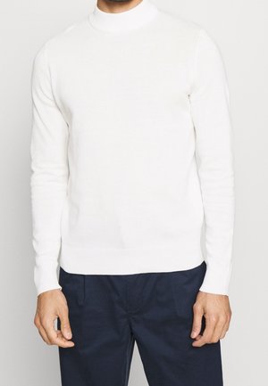 Mann trägt einen weißen langärmeligen Stehkragenpullover und dunkelblaue Hose, steht vor einem schlichten hellen Hintergrund.