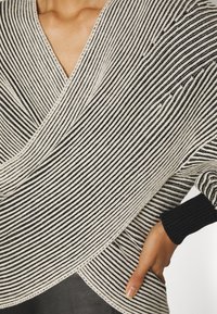 Gros plan d'une personne portant un pull croisé rayé noir et blanc avec des poignets noirs, main posée sur la hanche et encolure visible.