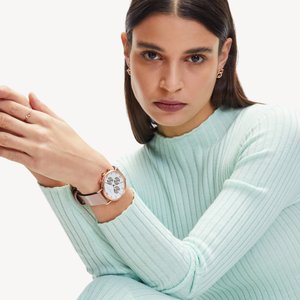 Montre ronde avec un cadran blanc, des accents en or rose et un bracelet rose clair. Le modèle porte un pull côtelé vert menthe à manches longues.