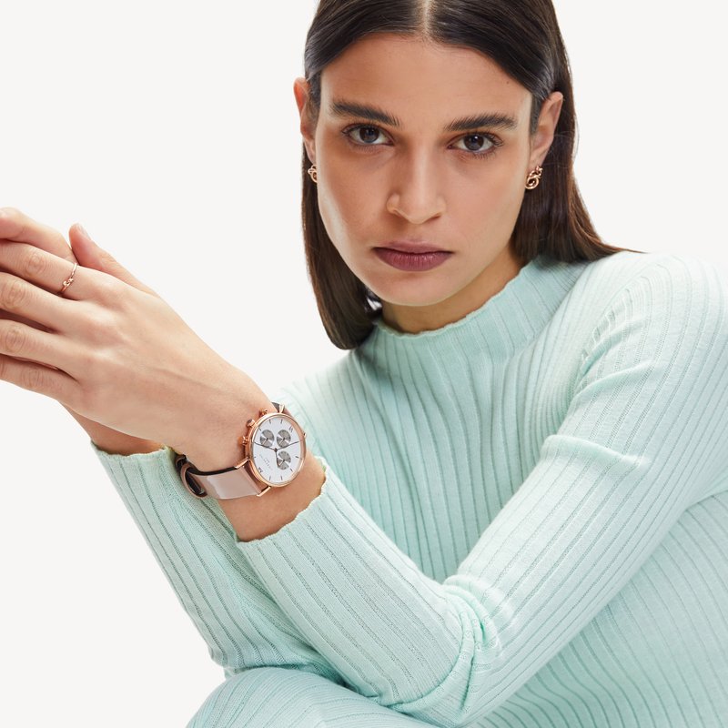 Runde Uhr mit einem weißen Ziffernblatt, roségoldenen Akzenten und einem hellrosa Armband. Das Modell trägt einen gerippten, mintgrünen Pullover mit langen Ärmeln.