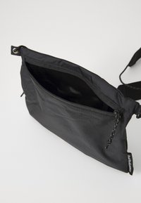 Sac en nylon noir à bandoulière avec une poche zippée, texture lisse et sangle réglable ; présente une étiquette de marque sur le côté.