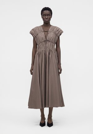 Femeie care poartă o rochie taupe fără mâneci, cu detalii adunate și șnururi, stând în fața unui fundal alb simplu.