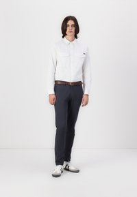 Chemise blanche à manches longues avec deux poches poitrine, pantalon slim-fit marine et baskets blanches avec des accents noirs. Le modèle se tient devant un fond uni.