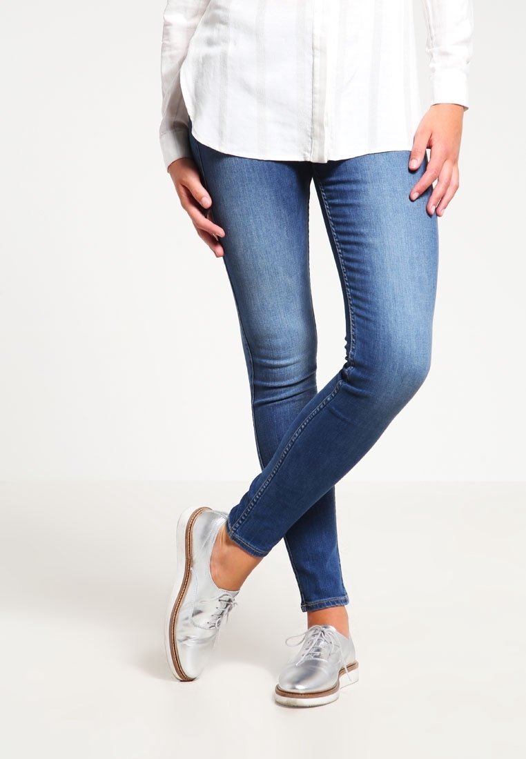Personne portant un jean skinny bleu, une chemise blanche boutonnée et des chaussures à lacets argentées métalliques, debout les jambes croisées.
