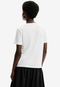 Camiseta de algodón blanca con cuello redondo, mangas cortas y corte holgado, vista desde la parte posterior. Textura suave con detalles mínimos.