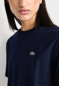 T-shirt en coton bleu marine avec col rond et manches courtes ; arbore un petit logo de crocodile vert brodé sur la poitrine.