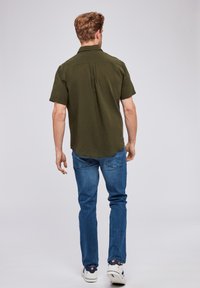 Camicetta verde oliva a maniche corte con orlo dritto, spacco posteriore e tessuto testurizzato, abbinata a jeans blu e sneakers bianche.