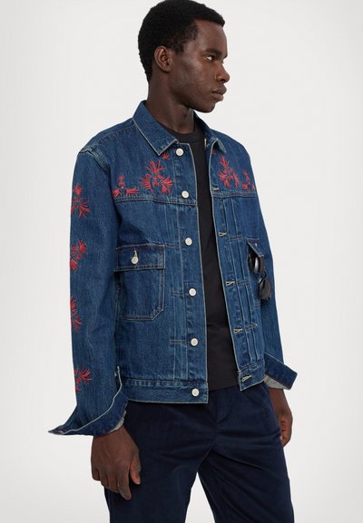 Denim jacket featuring red floral embroidery, light wash, traditional collar, front pockets, and button closure. Worn over a black shirt.