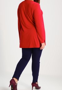 Röd långärmad topp med rundad och krökt söm bak, kombinerad med marinblå slim-fit byxor och burgundy högklackade skor.