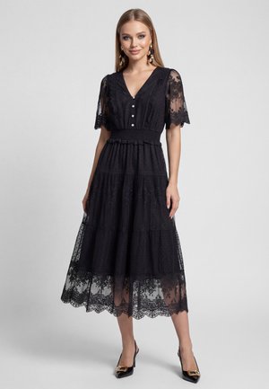 Donna che indossa un vestito midi in pizzo nero con maniche corte, corpetto abbottonato, vita arricciata e orlo smerlato, abbinato a tacchi neri.