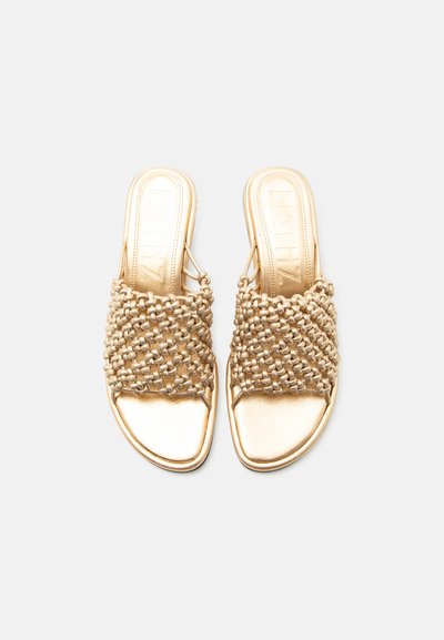 Arany színű, nyitott orrú, papucs típusú szandálok párosa, fonott pántokkal és lapos talppal, fehér háttéren.