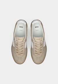 Beige suede sneakers med hvite skinnakenter, rund tå, flate skolisser og "BOSS"-merke på tungen og siden. Gummisåle.