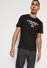 Crna pamučna majica s istaknutim bijelim logom "TOMMY JEANS" na prsima. Uparena s tamnim trapericama koje prikazuju ponovljeni uzorak loga.