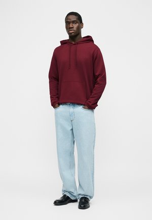 Mann steht mit den Händen in den Taschen, trägt einen burgunderroten Hoodie, hellblaue Jeans mit weitem Bein und schwarze Lederschuhe vor einem schlichten weißen Hintergrund.