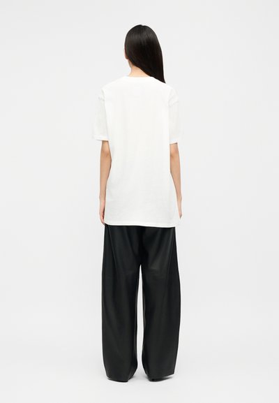 Hosszú sötét hajú személy, fehér bő szabású rövid ujjú inget és széles fekete nadrágot viselve, háttal áll egy egyszínű fehér háttér előtt.