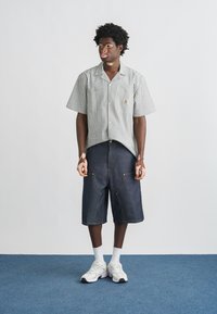 Νεαρός άνδρας με λεύκη φοράει ριγέ κοντομάνικο πουκάμισο, μακριά τζιν σορτς, λευκές κάλτσες και αθλητικά παπούτσια σε μπλε χαλί.