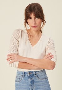 Sweter w serek w jasnym beżu z teksturowanymi, wzorzystymi rękawami z siateczki. Tkanina ma miękkie wykończenie; zestawiony z niebieskimi spodniami o wysokim stanie.