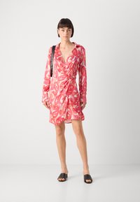 Robe portefeuille à motif paisley rose avec des manches longues, un décolleté en V et un détail de taille nouée. Des mules noires à bout ouvert complètent le look.