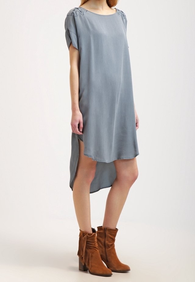 Šedé šaty s krátkým rukávem a zakřiveným lemem a bočními rozparky. Vyrobené z hladkého materiálu. Spárováno s hnědými kotníkovými botami s třásněmi.