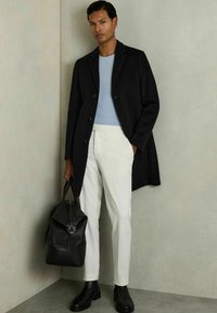 Palton lung negru, cămașă bleu deschis, pantaloni albi, cizme negre până la gleznă și o geantă din piele neagră cu fermoar. Fundal cu textură de perete.