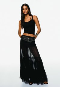 Black tank top paired with a sheer black maxi skirt featuring tiered layering and a studded black belt. Smooth texture and flowing design.