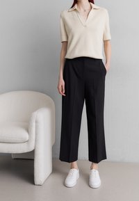 Béžový pletený polo triko s krátkým rukávem, kombinovaný s černými tailored kalhotami a bílými teniskami, stojící na texturované podlaze.