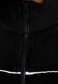 Schwarze Zip-Up-Jacke mit hohem Kragen, geripptem Material und einem weißen horizontalen Streifen über der Brust, ausgestattet mit einem Plastikreißverschluss.