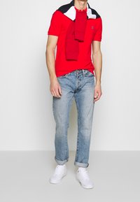 Röd polo skjorta med korta ärmar, vit logotyp; ljusblå jeans med avslappnad passform; vita sneakers; marinblå och röd tröja knuten avslappnat runt halsen.