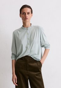 Blusa verde chiaro a maniche lunghe con colletto alto e apertura con bottoni, abbinata a pantaloni in velluto a coste marrone scuro, caratterizzati da una texture a coste verticali.