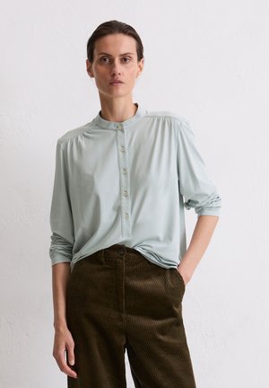 Lichtgroene blouse met lange mouwen, een staanderkraag en knoopsluiting, gecombineerd met donkerbruine corduroypants met verticale ribstructuur.
