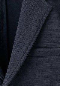 Blazer navy con tessuto strutturato, revers a incrocio e tasca sul petto singola. Il materiale appare liscio con dettagli di cuciture fini.