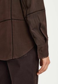 Chemise longue marron texturée à manches longues avec des coutures contrastées. Comprend des poignets à boutons et un ourlet arrondi, assortie à un pantalon noir.