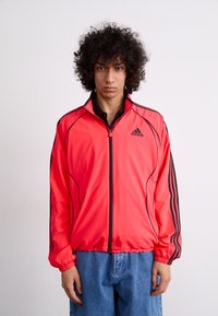 Bright red sports jacket with black accents, zip closure, and elastic cuffs. Features three black stripes along the sleeves. Lightweight fabric.