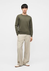 Άντρας που φοράει πουλόβερ μακρυμάνικο ελιάς, μπεζ παντελόνι φαρδύ και λευκά παπούτσια, στέκεται μπροστά σε απλό λευκό φόντο.