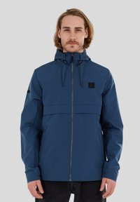Chaqueta impermeable azul con cierre de cremallera, capucha ajustable y dos bolsillos. Presenta un panel horizontal en el pecho y detalle del logo en negro.