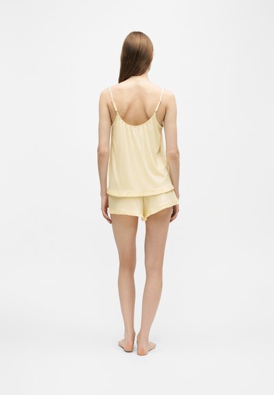 Femme debout pieds nus, de dos, portant un haut sans manches jaune pâle à bretelles réglables et un short assorti avec des bords volantés.
