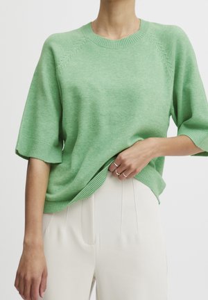 Personne portant un pull en maille à manches courtes vert clair et un pantalon crème taille haute, tenant l'ourlet du pull avec une main.
