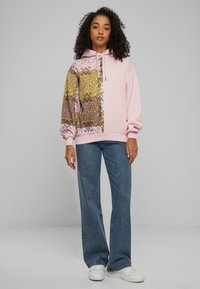 Hoodien har en rosa kropp med mønstrede gule og brune lapper, snorhette og ribbede mansjetter. Kombinert med brede jeans og sneakers.