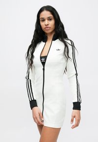 Λευκό φόρεμα με φερμουάρ και μαύρες λεπτομέρειες, με μακριά μανίκια, τρεις μαύρες ρίγες κατά μήκος κάθε μανικιού κ�αι μίνιμαλ λογότυπο Adidas στο στήθος.