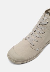 Beżowy trampki w stylu high-top z płóciennym materiałem, gumowym noskiem, sznurowanym przodem i teksturowaną podeszwą, ukazane z bliskiego, ukośnego widoku z góry.