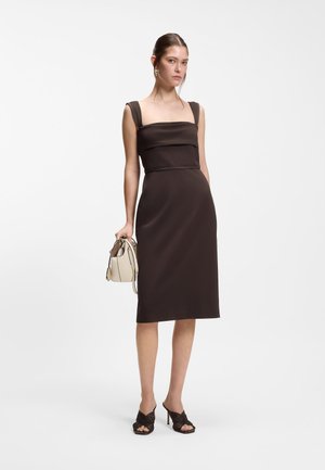 Kobieta ubrana w bezrękawnikową midi sukienkę w ciemnobrązowym kolorze, czarne sandały na obcasie, złote kolczyki, trzymająca małą beżową torebkę, stojąca na białym tle.