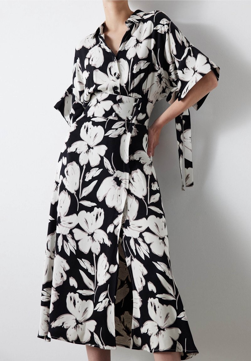 Femme portant une robe midi noire avec un grand imprimé floral blanc, manches mi-longues, col et fente devant, debout contre un mur blanc.