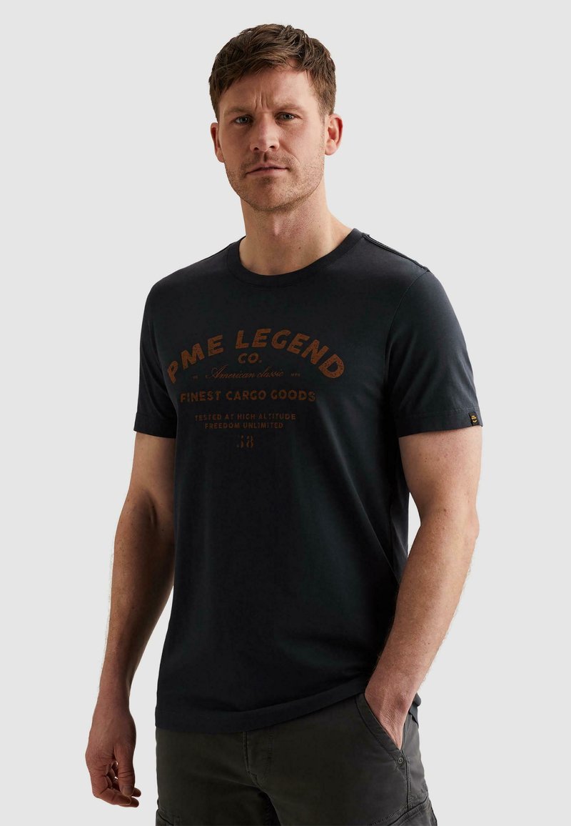 Mand iført sort PME Legend grafisk t-shirt og mørke cargobukser, stående med den ene hånd i lommen mod en ensfarvet lys baggrund.