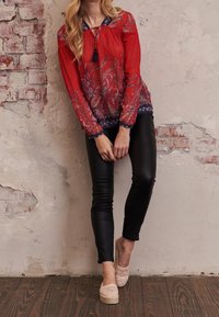 Röd mönstrad blus med paisleymönster, marinblå kant och knytdetalj vid halsringningen, kombinerad med svarta tajta byxor och beigefärgade espadrillos.